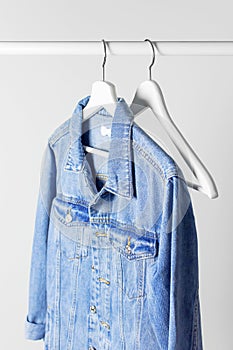 Blue denim jacket on white wooden coat hanger on a rod against light gray wall flat lay copy space. Denim, fashionable jacket,