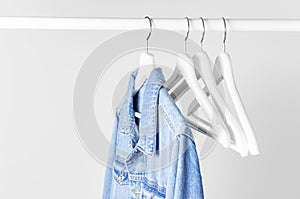 Blue denim jacket on white wooden coat hanger on a rod against light gray wall flat lay copy space. Denim, fashionable jacket,