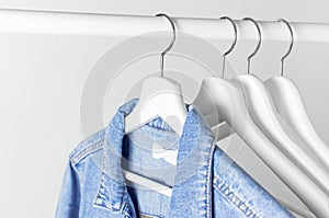 Blue denim jacket on white wooden coat hanger on a rod against light gray wall flat lay copy space. Denim, fashionable jacket,