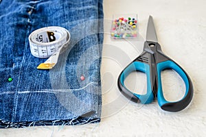 Blue denim shorts, roll of tailor tape with centimeters and inches, multi coloured headed sewing pins in a box and scissors.