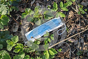 Blue disposable medical face mask between green plants and old dry leaves, thrown away like garbage on nature