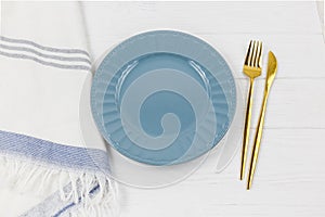 Blue empty plate with fork, knife, white striped textile napkins on wooden table. Golden cutlery, table setting utensils
