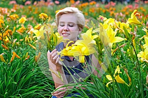 Blue-eyed blonde woman among blooming yellow lilies in the garden