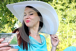 blue-eyed brunette in a turquoise suit and white hat looks coquettish in a small mirror. girl is looking at herself in small