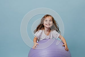 Blue-eyed curly toddler girl laughs playing on lilac fitness ball