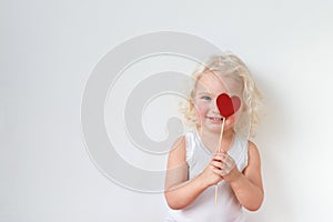 Blue eyed pretty small kid dressed casually, has fun indoors, covers eye with heart stick, glad to spend free time with