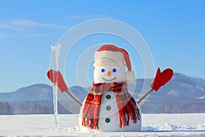 The blue eyed smiling snowman in red hat, gloves and plaid scarf holds the icicle in hand. Joyful cold winter morning.