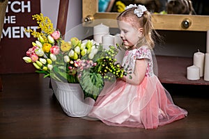 Blue-eyed sweet girl in a pink dress sitting near a vase with tulips, mimosa, berries and greens and smiling