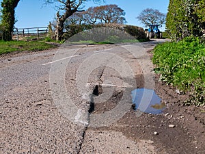 A blue farm tractor approaches surface damage to tarmac on a rural road is marked for repair with white spray paint.