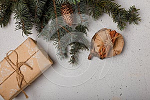 Blue fir tree branch with big cone, wrapped present box, wooden cut and cinnamon sticks on white wooden background. Christmas card