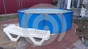 The blue frame swimming pool is located on an asphalt lot near the house. Next to it is a plastic sun lounger and the porch of the