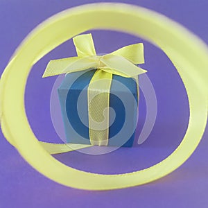 Blue gift box at the end of the spiral yellow ribbon,  purple background, Square.