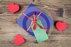 Blue gift box with red ribbon, blank gift tag and and red hearts on wooden background.