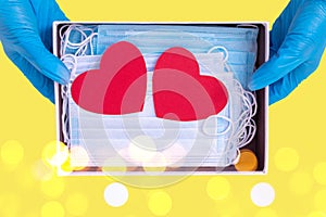 Blue gloved hands hold an open gift box with protective masks and two red heart shapes on the trendy Illuminating