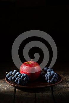 blue grapes and red apple in a clay plate on a wooden table
