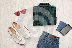 Blue-green checkered shirt, glasses, sneakers, jeans, phone and passport. Wooden background. Fashionable concept