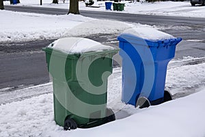 A blue and a green trash can covered with snow by the side of a snow covered, freshly plowed street waiting for garbage pickup