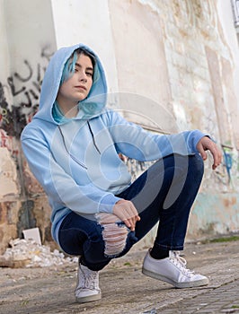 Blue haired Teenage girl in light blue oversize hoodie sitting on her haunches near graffiti wall