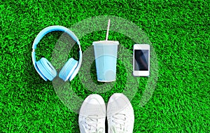 Blue headphones, white smartphone, cup of fruit juice with sports sneakers shoes on a green grass textured background