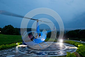 A blue helicopter is parked on a private helipad against a blue evening.