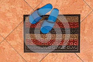Blue homemade slippers stand on the foot carpet against the background of the floor tiles, the top view from above