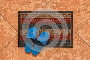 Blue homemade slippers stand on the foot carpet against the background of the floor tiles, the view from above