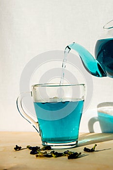 Blue hot tea is poured into a glass cup. A teapot with blue Thai tea, which is poured into a mug against a white wall.