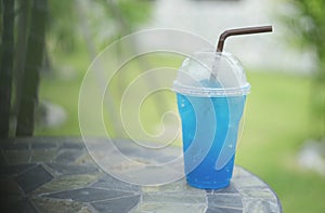 Blue italian soda in takeaway cup on marble stone table with blurred green grass and bamboo in background,selective focus