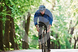 In blue jacket. Cyclist on a bike is on the asphalt road in the forest at sunny day