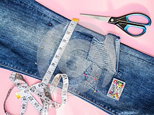 Blue jeans with large hole folded in half, sewing pins in a box, white tailor tape with centimeters and inches and scissors.