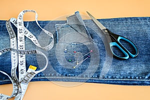Blue jeans with a large hole on a pant leg below the knee, multi coloured headed sewing pins, tailor tape and scissors