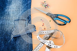 Blue jeans with a large hole on a pant leg below the knee, sewing pins, tailor tape and scissors