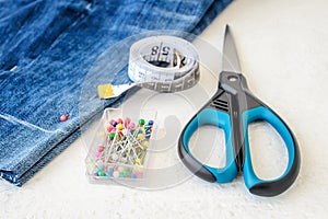 Blue jeans shorts, roll of tailor tape with centimeters and inches, multi coloured headed sewing pins in a box and scissors.