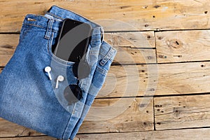 Blue jeans with smartphone, earphones and glasses lying on wooden surface