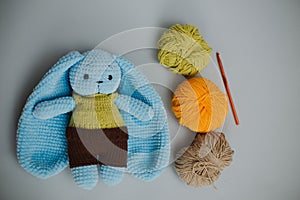 blue knitted rabbit with big ears in a yellow vest and brown pants with three skeins of yarn, and knitting needles