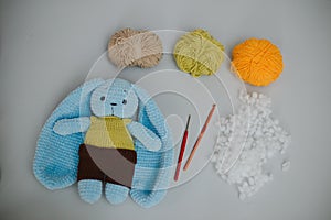 blue knitted rabbit with big ears in a yellow vest and brown pants with three skeins of yarn, and knitting needles