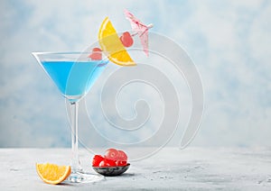 Blue lagoon summer cocktail in martini glass with sweet cocktail cherries and orange slice with umbrella on blue background