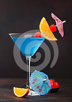 Blue lagoon summer cocktail in martini glass with sweet cocktail cherries and orange slice with umbrella on dark background