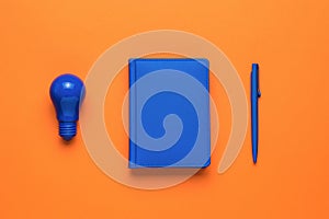 A blue light bulb, a blue notebook and a blue pen on an orange background. Flat lay