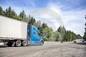 Blue long haul big rig industrial semi truck with dry van semi trailer running on the long divided highway road