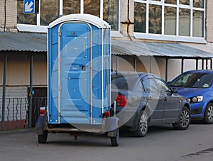 A blue mobile toilet stands on a small trailer of a passenger car