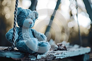 Blue Monday concept. Old blue toy teddy bear sitting on old wooden swing outdoors. Feelings of depression, sadness