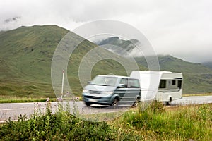 A blue MPV car with a trailer caravan driving on a road in the landscape of Scottish Highlands in misty weather with motion blur