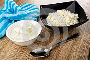 Blue napkin, bowl with sour cream, black bowl with defatted cottage cheese, spoon on wooden table