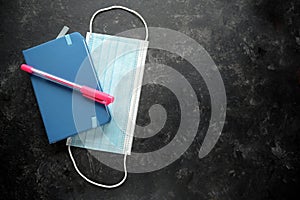 Blue notebook, pink pen and a medical protection face mask against coronavirus and covid 19 infection, dark slate background with