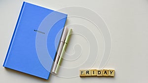 Blue notebook with two pens and wooden word `Friday` on a beige paper background. Top view. Close-up.