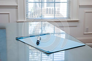 A blue notepad with a pen sitting on a glass top table near a window in a business ready for client notes and notetaking
