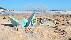 Blue paper boat and three dollar paper bills half in sand on beach near sea.
