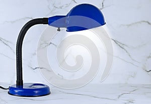 A blue personal sized desk lamp on a marble desk in front of a marble wall.