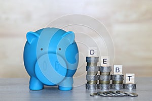 Blue piggy bank and stack of coins money with wooden blocks with the word debt on wooder background, Debt and risk management